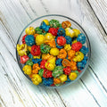 Bowl of colorful Galaxy Popcorn featuring blue, green, red, orange, and yellow fruit-flavored popcorn on a white wood surface.