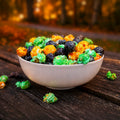 A white ceramic bowl filled with green, orange, and black Halloween Mix popcorn on a rustic dark wooden table, with a blurred autumn forest background.